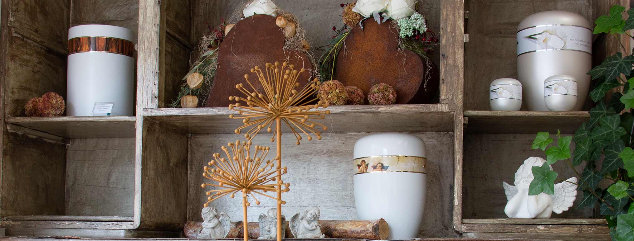 Ein rustikales Regal mit verschiedenen stilvollen Urnen, darunter schlichte weiße Modelle sowie Urnen mit Blumendekoration, umgeben von Trockenblumen und Engelsfiguren.