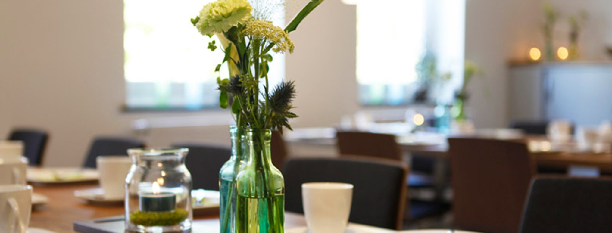Eine gedeckte Kaffeetafel mit dezenten grünen Glasvasen, frischen Blumen und Kerzen in einem hellen Raum, der für eine Trauerfeier oder einen Erinnerungskaffee nach der Beisetzung vorbereitet ist.